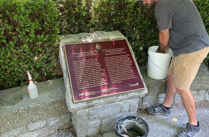 A man leaning down to wash a bronze Japanese Canadian Soldiers historical plaque set in stone outdoors.