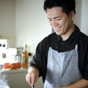 Asian man cooking in black shirt and grey apron with a smile.