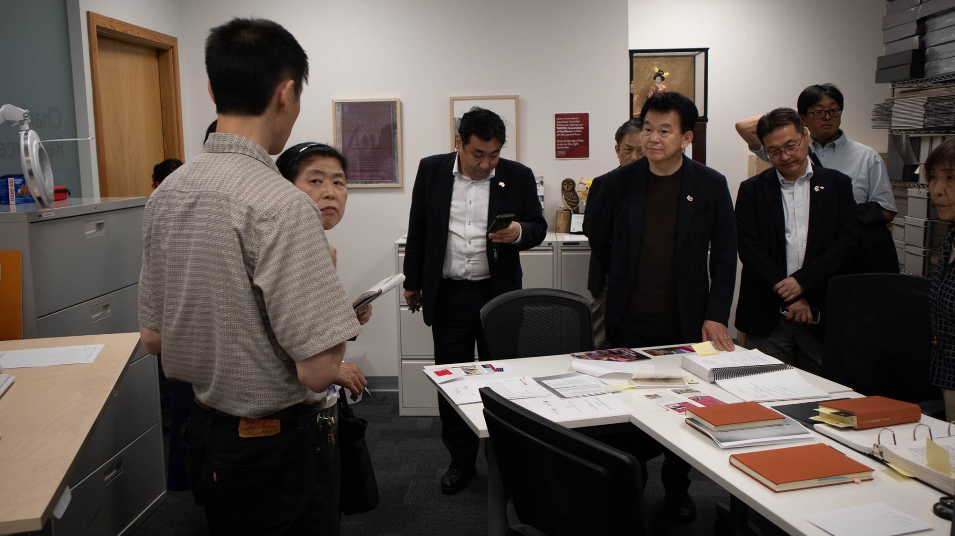 A group of visitors in suits and casual wear gathered around a white office table covered with documents, notebooks, and pamphlets during an informational briefing inside an archival room.