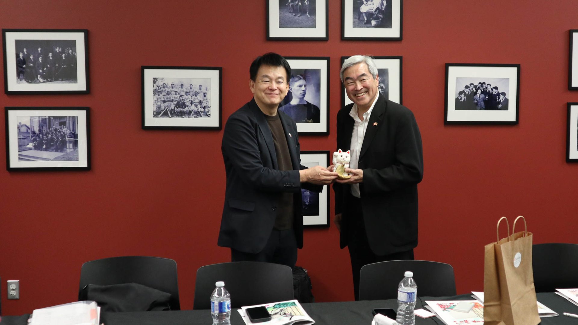 Two men in dark suits smiling in front of a deep red wall with framed black-and-white historical photos. The man on the left is presenting a small white Beckoning Cat (Maneki-neko) figurine to the man on the right.