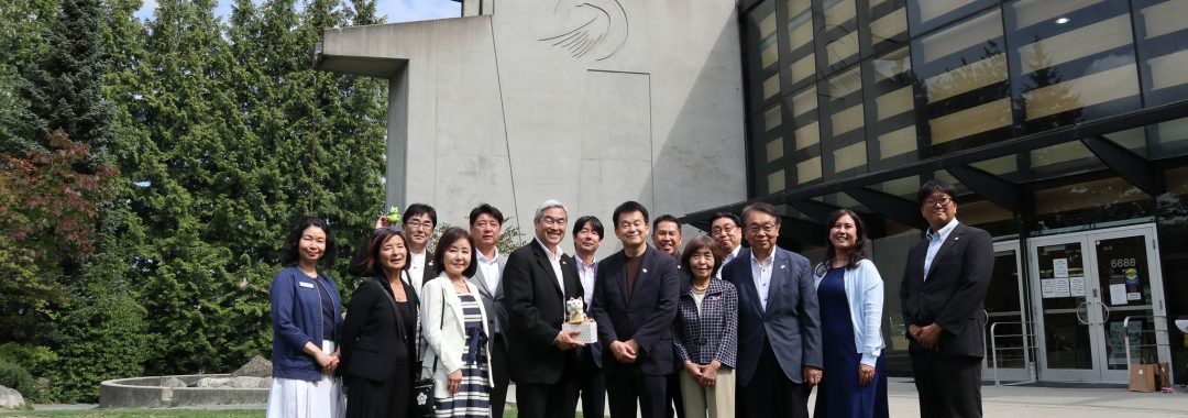 A group pose outdoors on a green lawn in front of a modern concrete building with a large glass facade. The group of men and women, dressed in business attire, stand side-by-side while a man near the center holds a small gift item.
