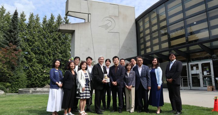 A group pose outdoors on a green lawn in front of a modern concrete building with a large glass facade. The group of men and women, dressed in business attire, stand side-by-side while a man near the center holds a small gift item.