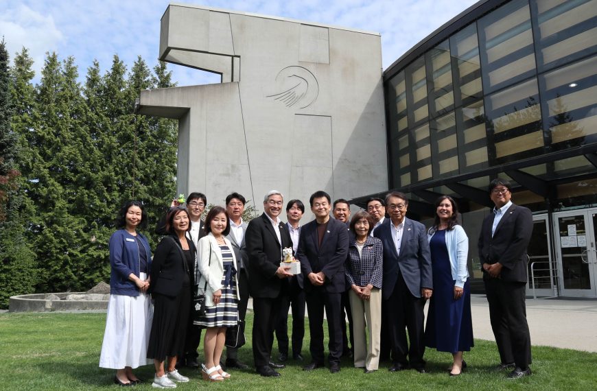 A group pose outdoors on a green lawn in front of a modern concrete building with a large glass facade. The group of men and women, dressed in business attire, stand side-by-side while a man near the center holds a small gift item.