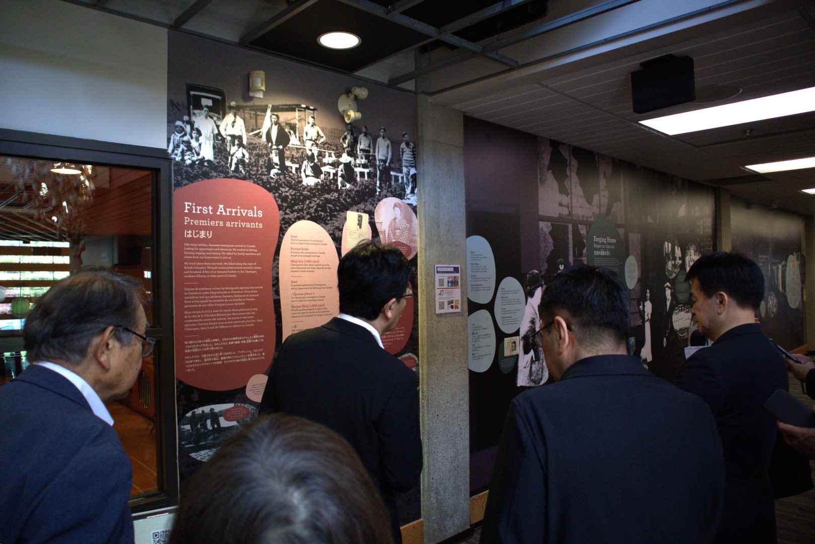 A group of visitors in dark suits listening intently while looking at a large wall exhibit titled "First Arrivals", which features historical photographs, red and black text panels, and circular information graphic displays about Japanese immigrants in Canada.
