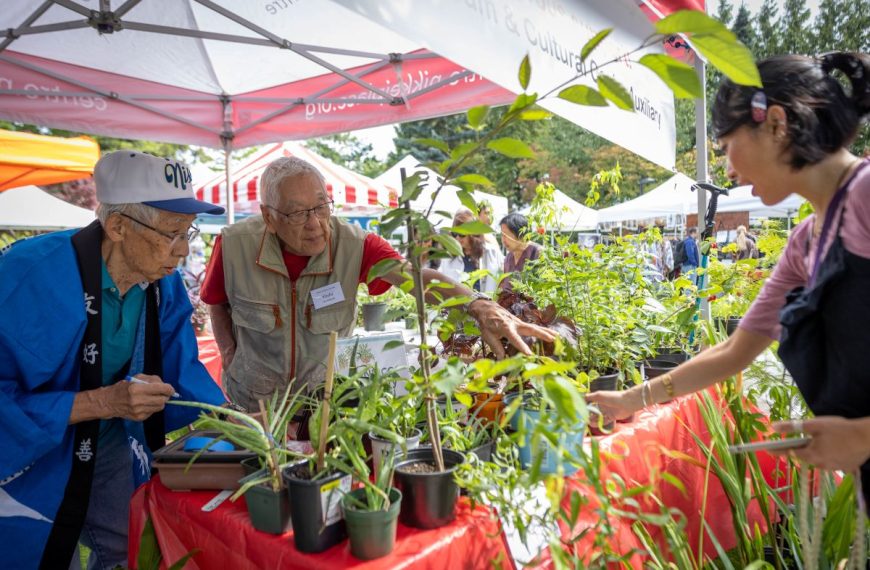Two senior men inspect potted plants for sale at an outdoor community garden market booth under a white canopy tent.