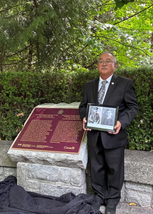 David Mitsui standing next to a historic commemorative plaque, holding a framed portrait photo.