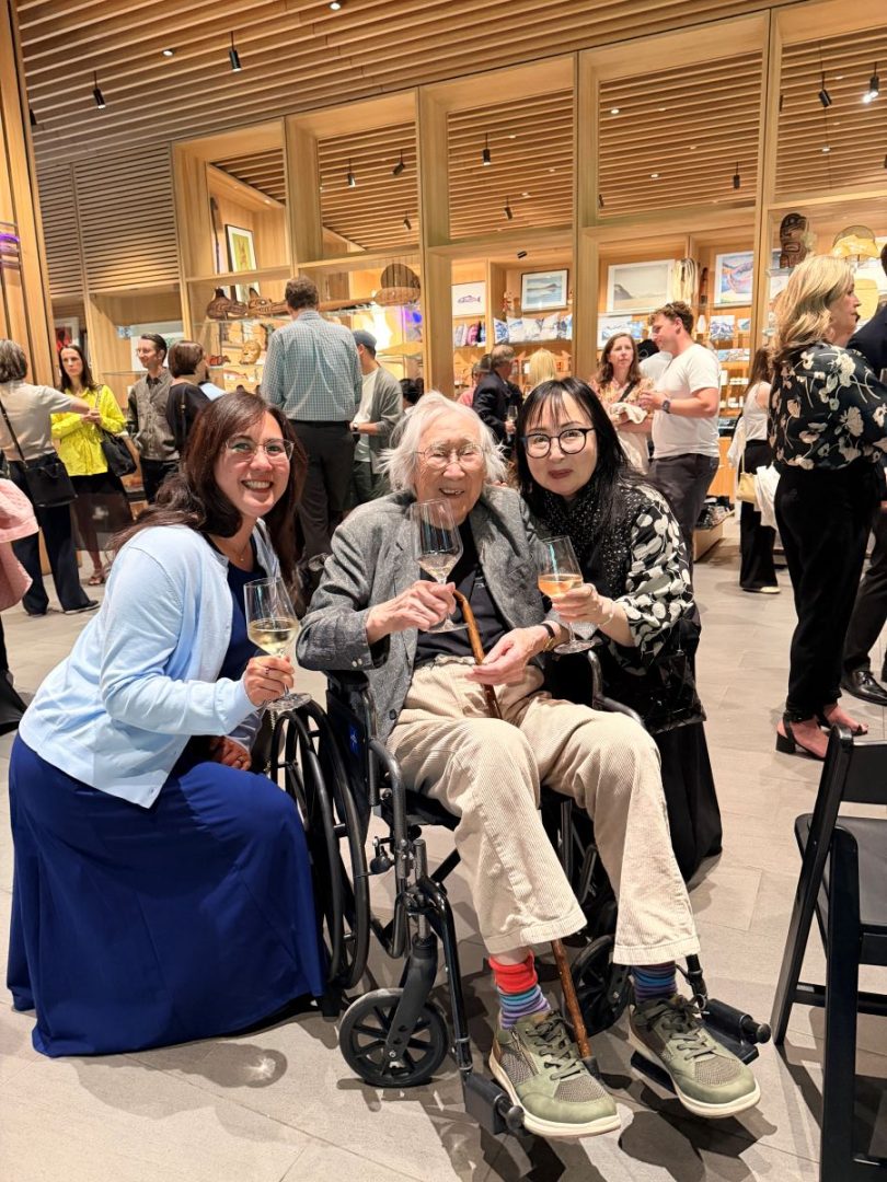 Tak Tanabe seated in a wheelchair alongside Sherri and Karah, all smiling and holding glasses of wine at an indoor event.