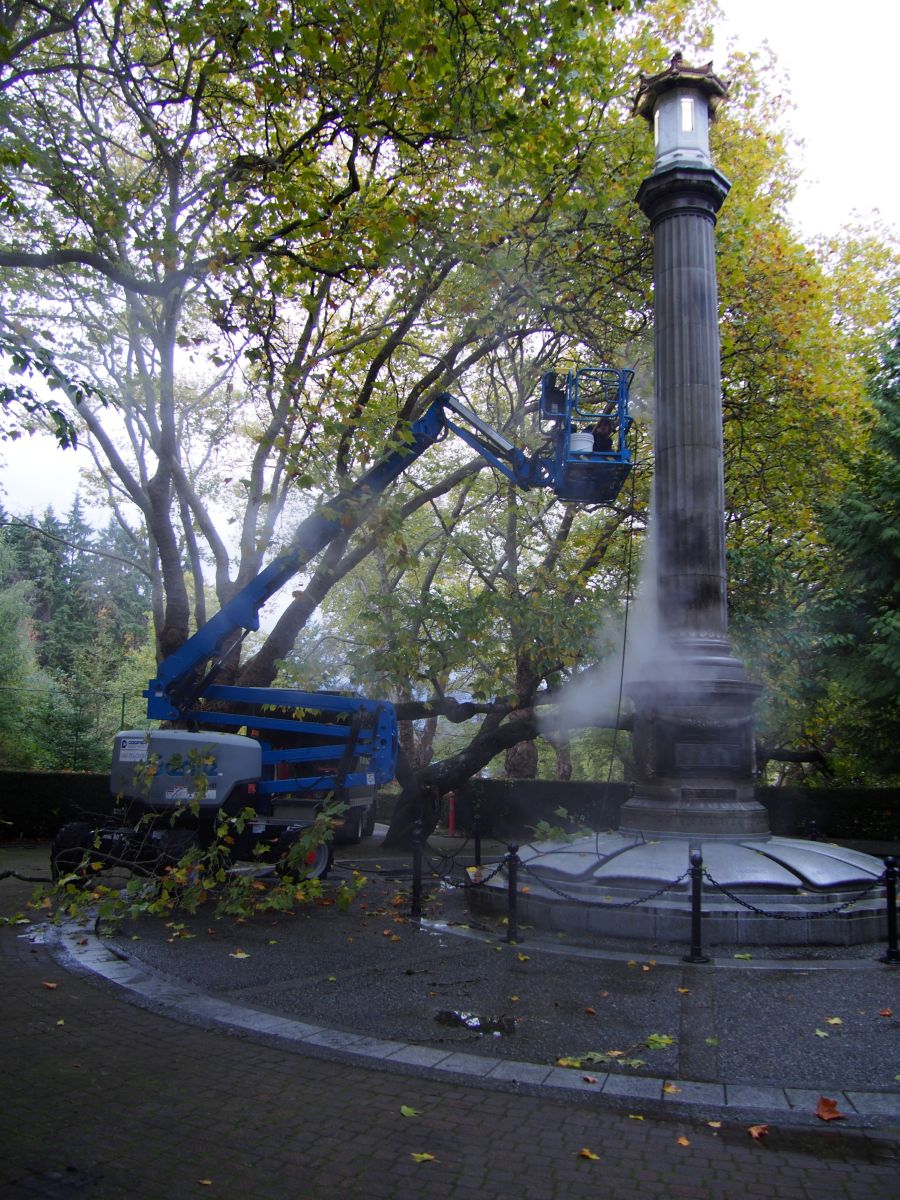 A boom lift pressure washing the stone Japanese Canadian War Memorial cenotaph surrounded by trees.