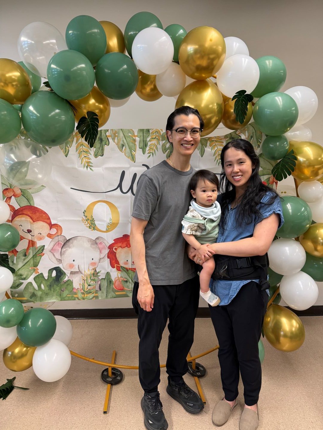 A smiling family of three posing in front of a green, white, and gold balloon arch at an event with animal-themed backdrop decorations.